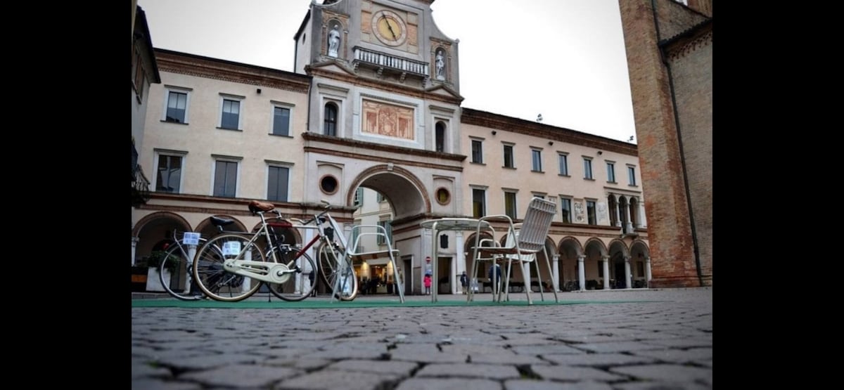 A cobblestone square is depicted, featuring a historic building with an ornate clock and archways. Two bicycles are parked near a set of metal chairs and a table, inviting visitors to enjoy the ambiance of the central plaza.