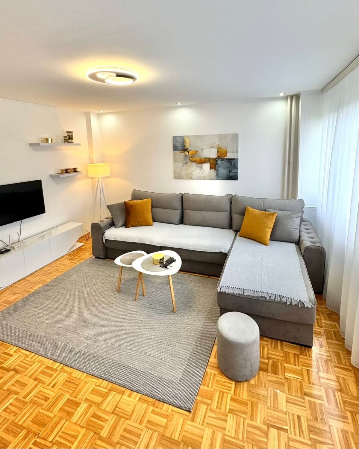 A modern living room features a comfortable grey sectional sofa adorned with two yellow accent pillows. A small round coffee table with two nested tables sits on a textured grey rug. A flat-screen TV is mounted on the wall, with soft lighting provided by a floor lamp.