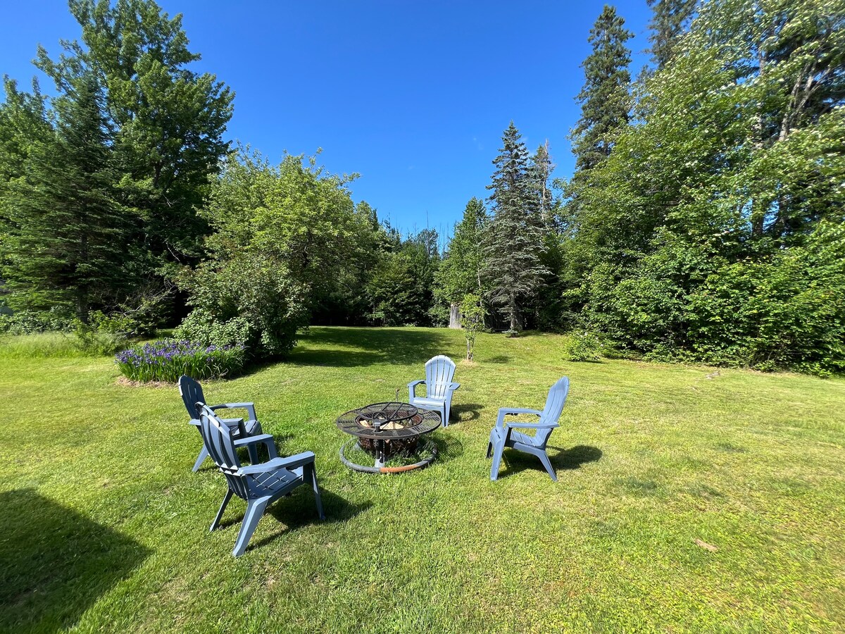 A spacious backyard features a fire pit surrounded by four blue Adirondack chairs, set against a backdrop of lush green grass and various trees. The area is illuminated by natural light, offering a peaceful space for outdoor gatherings.