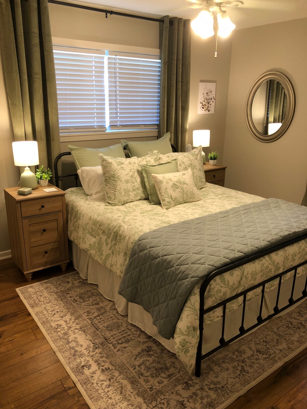 A well-appointed bedroom features a stylish metal bed with soft linens in soothing hues. Two bedside tables are graced with lamps and decorative plants. Natural light filters through the window's blinds, while a round mirror reflects the room's inviting décor, complemented by a decorative area rug.