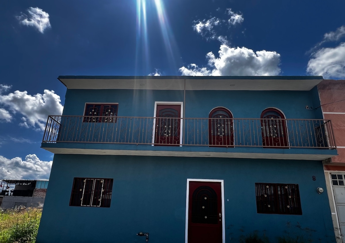 A two-story building is presented, featuring a vibrant blue exterior. Red doors and windows provide a striking contrast. A balcony extends from the second floor, allowing for views of the surrounding area. Fluffy clouds float in a clear blue sky above.