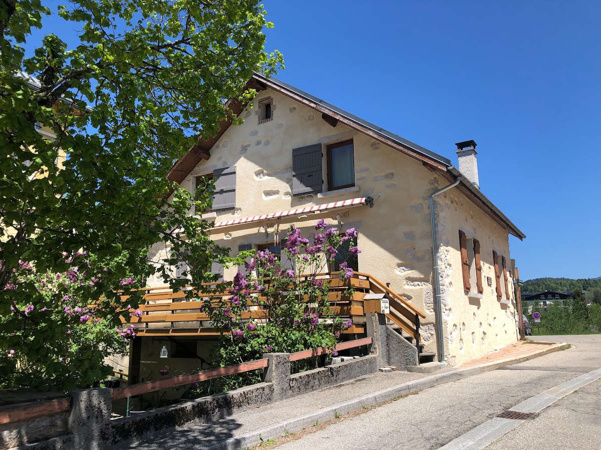 A charming stone house is viewed from the street, showcasing a light-colored facade with wooden shutters. A wooden balcony with a railing is adorned by blooming flowers, adding a touch of nature. The landscape features a clear blue sky, enhancing the welcoming appearance of the home.
