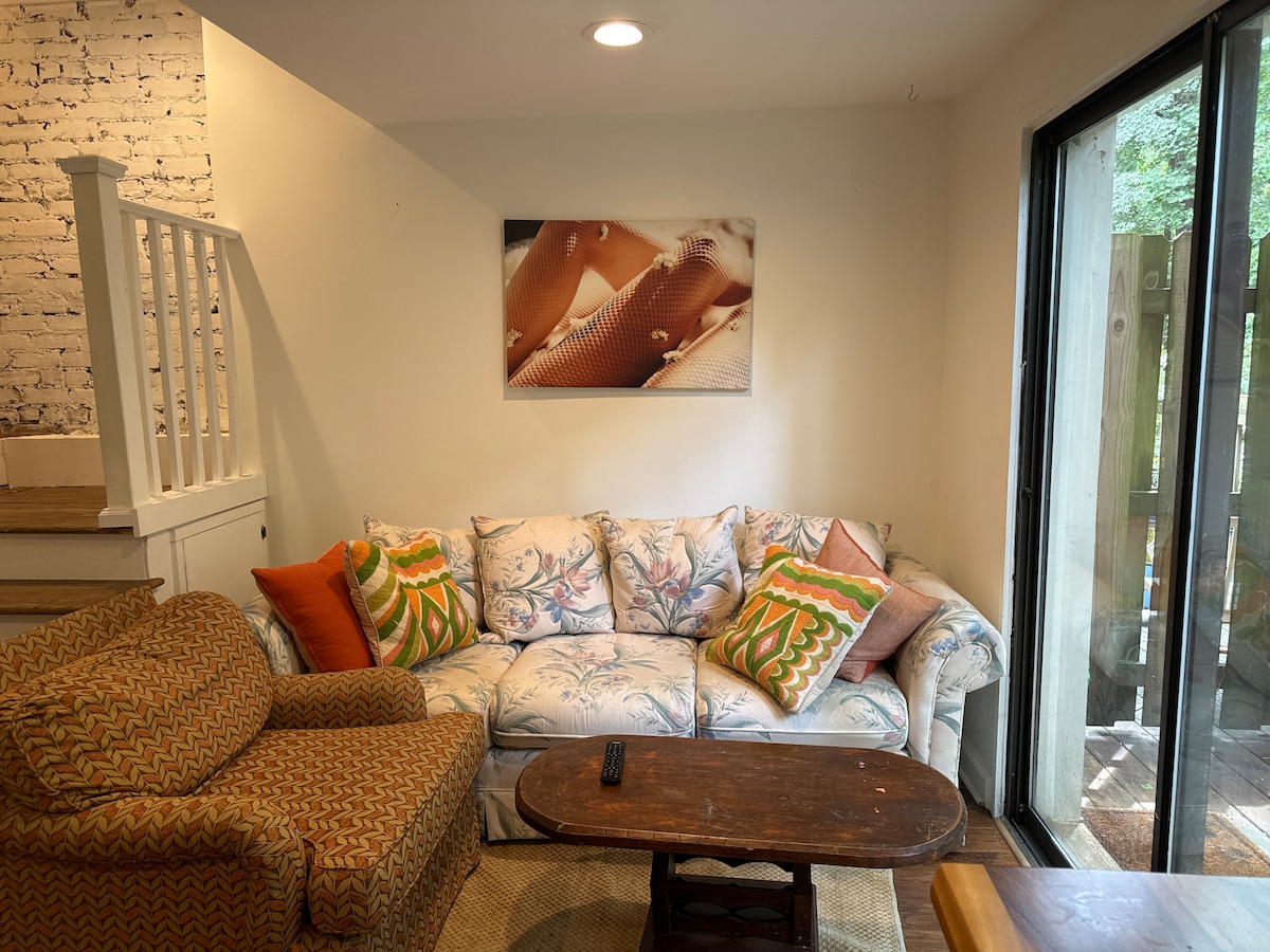 A comfortable living area features a floral-patterned sofa and an accent chair with a geometric design. A wooden coffee table occupies the center space, complementing the light wood tones of the room. Large glass doors allow natural light to fill the space and offer access to the outdoor area.