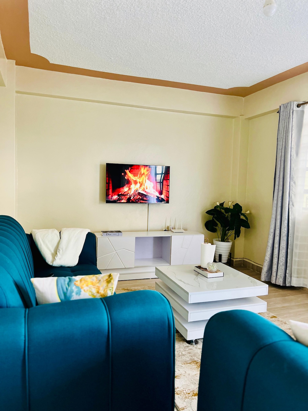 A modern living area features a teal sofa and a white coffee table. A flat-screen TV displays a flickering fire, creating a calming focal point. A potted plant adds a touch of greenery, while soft natural light enters through sheer curtains.