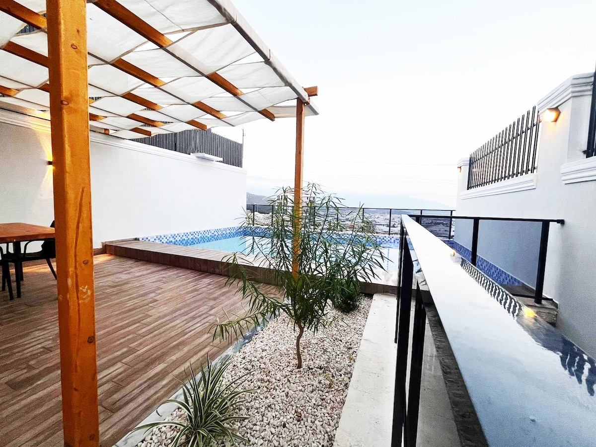 An outdoor area features a wooden pergola providing shade over a dining table. A private pool is visible, surrounded by a stone border and a landscaped garden. The distant horizon is evident, contributing to a sense of openness.