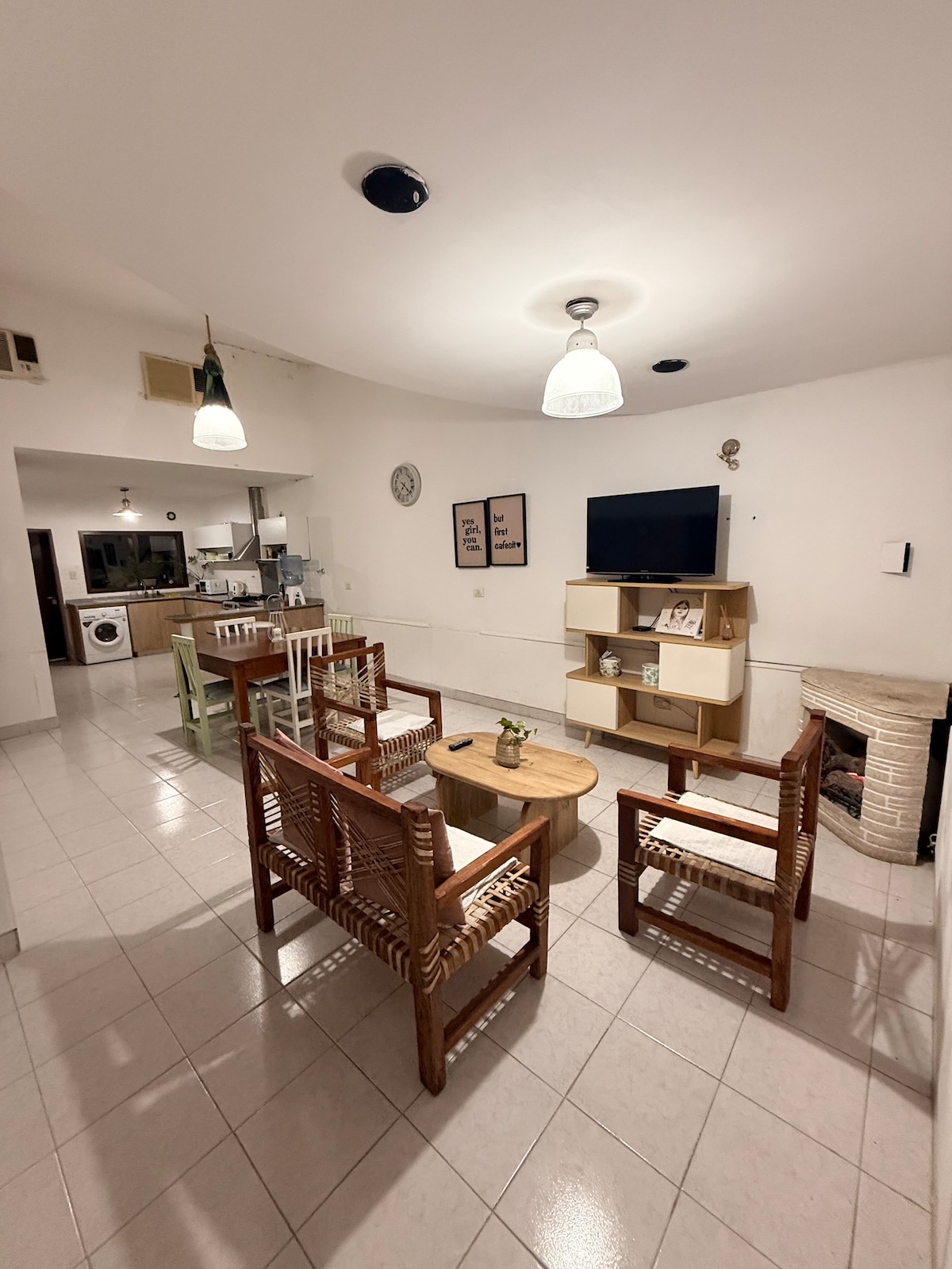 A living area features a set of wooden chairs arranged around a round coffee table. A television is mounted on the wall, and light fixtures hang from the ceiling. The space connects to a kitchen area, visible in the background.