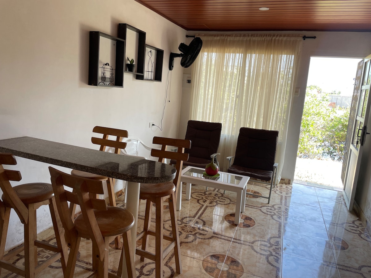 A light-filled living area is furnished with two comfortable chairs and a small coffee table. A bar with stools made of natural wood is visible alongside the window, which is draped with sheer curtains. The tiled floor features an intricate pattern that adds character to the space.