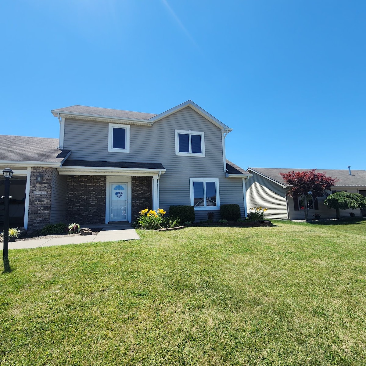 The exterior of a two-story home is shown, featuring a welcoming entrance and a well-maintained front yard with green grass. Flower beds add a touch of color, while the clear blue sky enhances the inviting appearance of the property.
