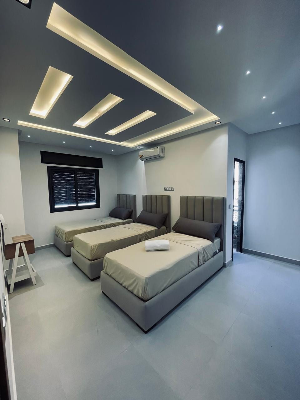 The sleeping area features two spacious beds with neutral-toned bedding. Indirect lighting along the ceiling highlights the modern design. A small side table is positioned next to one bed, and a window offers natural light, complemented by dark window coverings.