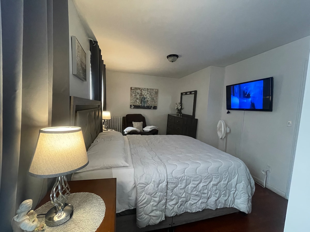 A well-furnished bedroom is presented, featuring a spacious bed dressed in white linens and a decorative headboard. A bedside lamp illuminates the room, which also includes a television mounted on the wall, dark curtains, and a comfortable seating area with an accent chair.