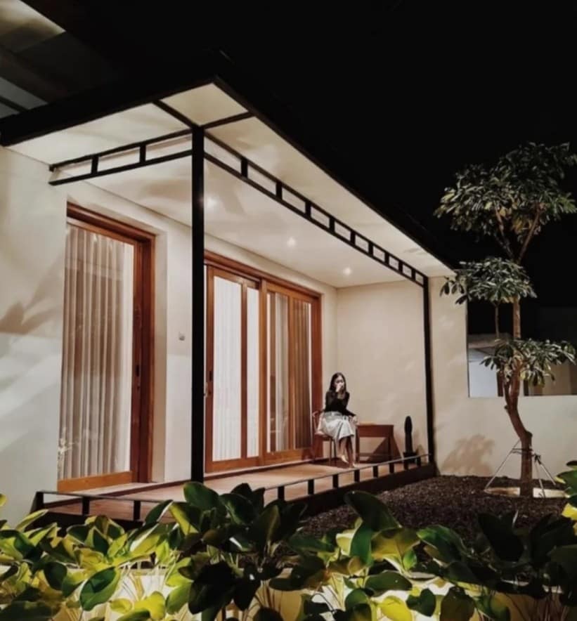 The exterior of a modern single-story house is viewed at night, featuring large glass doors framed by wooden accents. A serene statue sits on the porch, surrounded by lush greenery. Soft lighting highlights the minimalist design and architectural details.