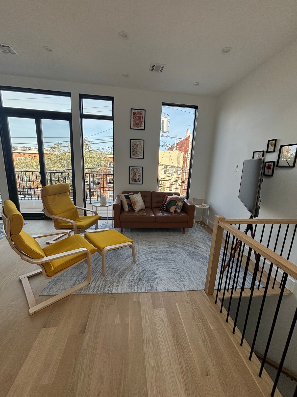 A bright living area features large windows allowing ample natural light. Two bright yellow lounge chairs are positioned near a brown leather sofa, creating a cozy seating arrangement. A light area rug lies beneath, while framed artwork adorns the walls.