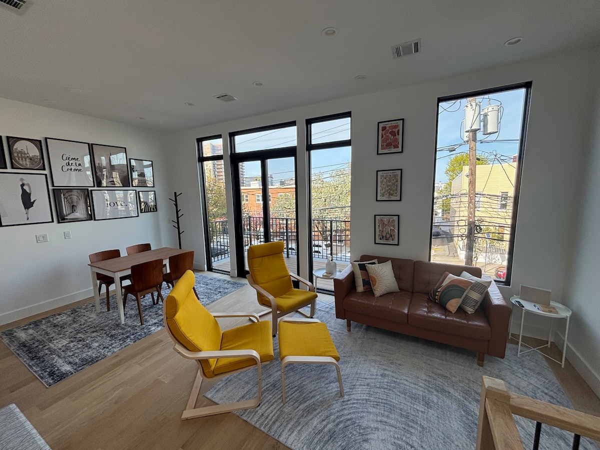 A bright and airy living space featuring large windows that provide ample natural light. Two yellow chairs are positioned near a light wood coffee table, alongside a brown leather sofa. A dining table for four is visible in the background, complemented by framed wall art.