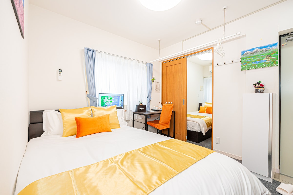 The spacious room features a large bed dressed in yellow and white linens. A desk and chair are present, along with a television visible in the corner. Natural light filters through sheer curtains, and a smaller bed can be seen in the adjacent area.