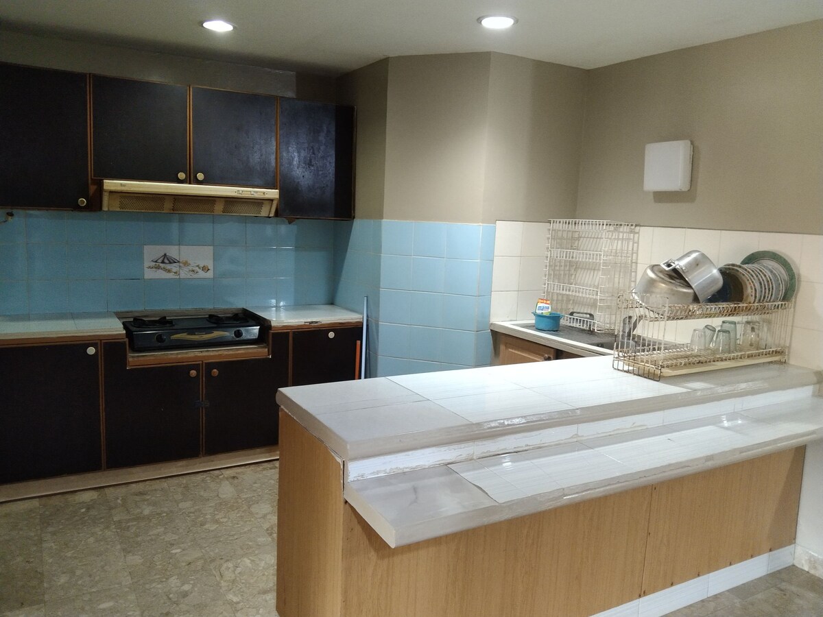 A functional kitchen features dark cabinetry complemented by light blue tiled walls. A countertop provides ample space for preparation, while a dish rack and cooking tools are neatly arranged. Under-cabinet lighting enhances the workspace, creating a bright and practical environment.