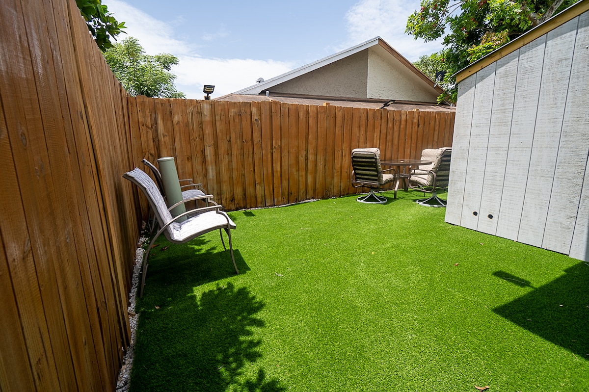 A spacious private backyard is visible, featuring a small grassy area with artificial turf. Two metal chairs and a round table are positioned on the grass, surrounded by wooden fencing and a storage shed in the background.
