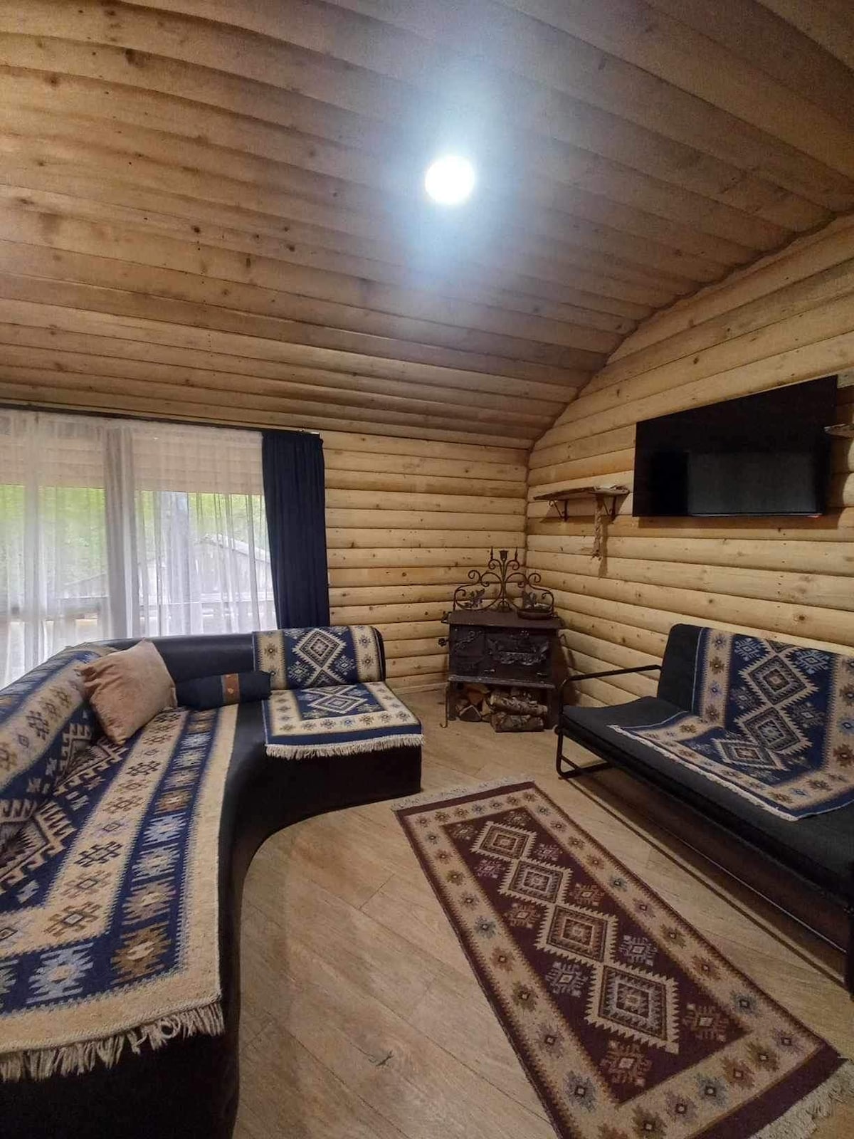 A cozy living area is highlighted by wooden walls and a ceiling featuring exposed beams. A dark sofa with decorative blankets faces a sleek television mounted on the wall. Light filters through large windows, enhancing the warm ambiance.