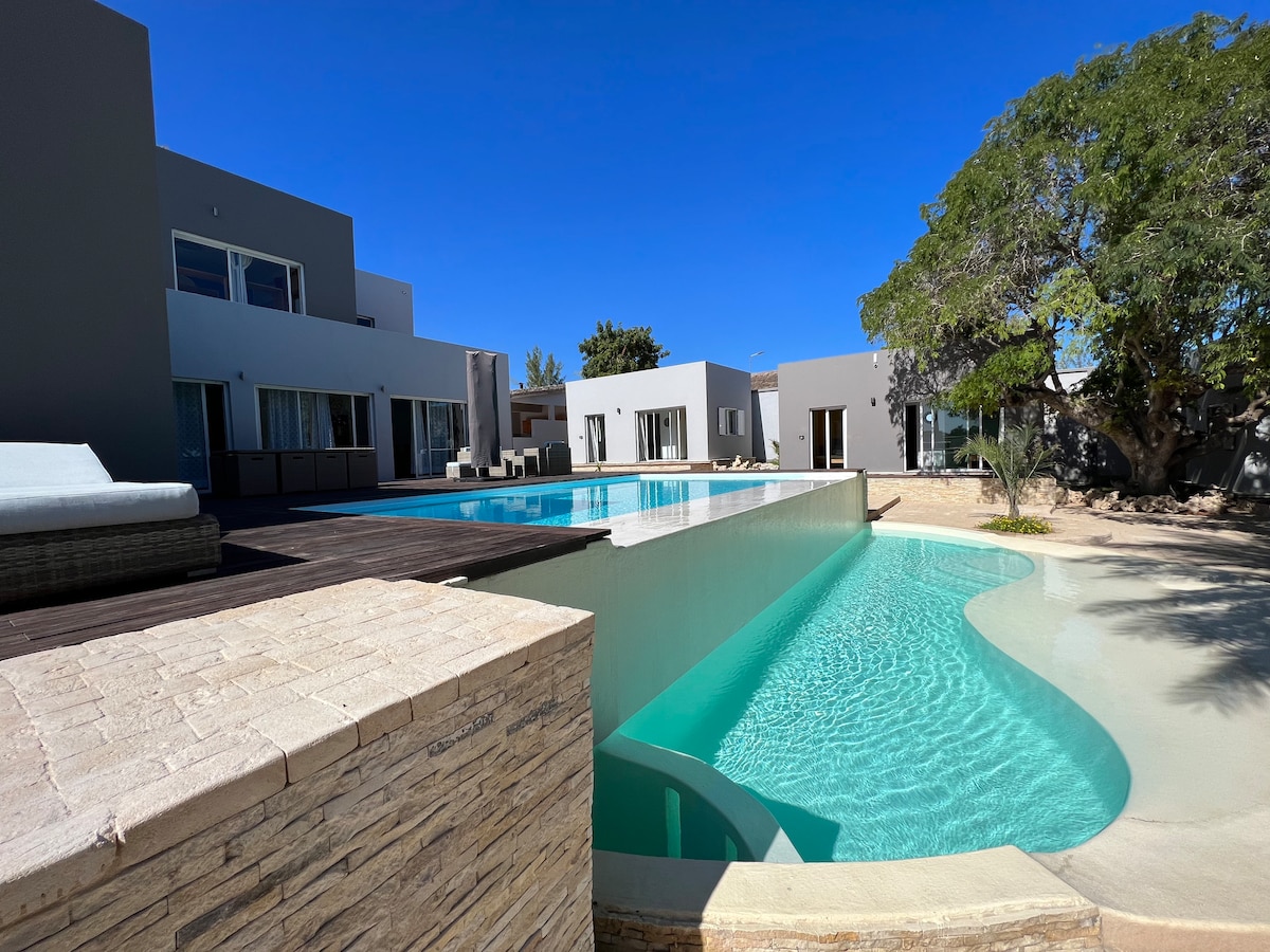 A modern villa is depicted featuring an inviting infinity pool that connects to a shallower pool for children. The property is surrounded by landscaped areas and mature trees, providing a tranquil outdoor environment. The sleek architecture reflects contemporary design with large windows and spacious decks.