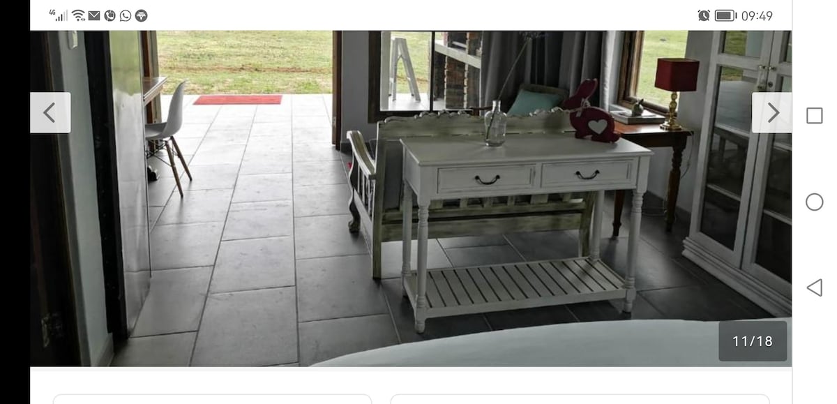 An elegant table with a drawer and a lower shelf sits in a well-lit space. A decorative item rests on the table, while a plush red lamp provides soft illumination nearby. Sliding glass doors open to a grassy outdoor area, enhancing the room's airy feel.
