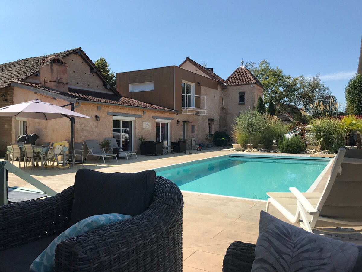 A large outdoor swimming pool is surrounded by sun loungers and patio furniture, creating a relaxing space. The building features a mix of rustic and contemporary architecture, with large windows and a flat roof. Lush greenery is visible in the background.