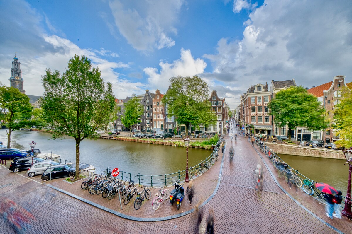 A scenic view captures the bustling Prinsengracht with historic buildings lining the water's edge. Bicycles are parked along the canal, while pedestrians pass over a charming bridge. Lush trees add greenery to the landscape, set against a dynamic sky with wispy clouds.