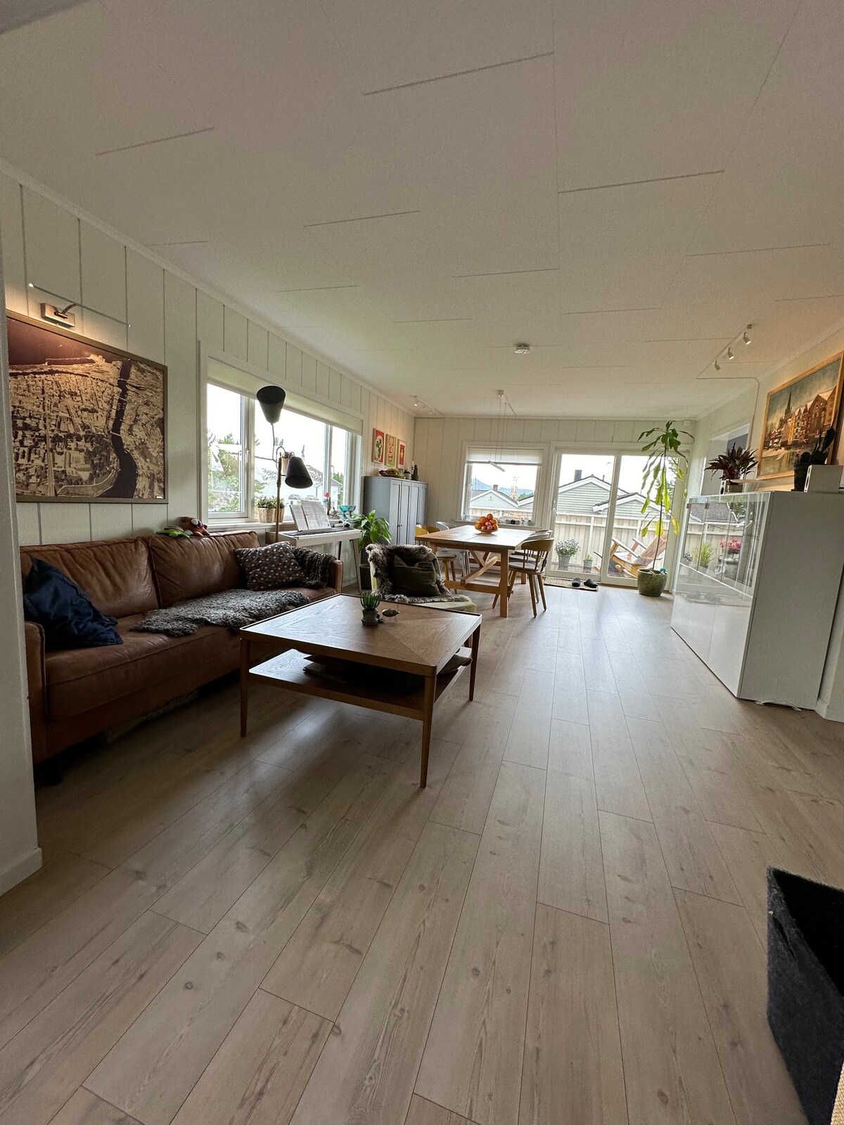 A bright and airy living space features a comfortable brown sofa adorned with decorative pillows. A wooden coffee table sits in front of the sofa, while large windows allow natural light to fill the room. A dining area is visible in the background, accompanied by indoor plants.