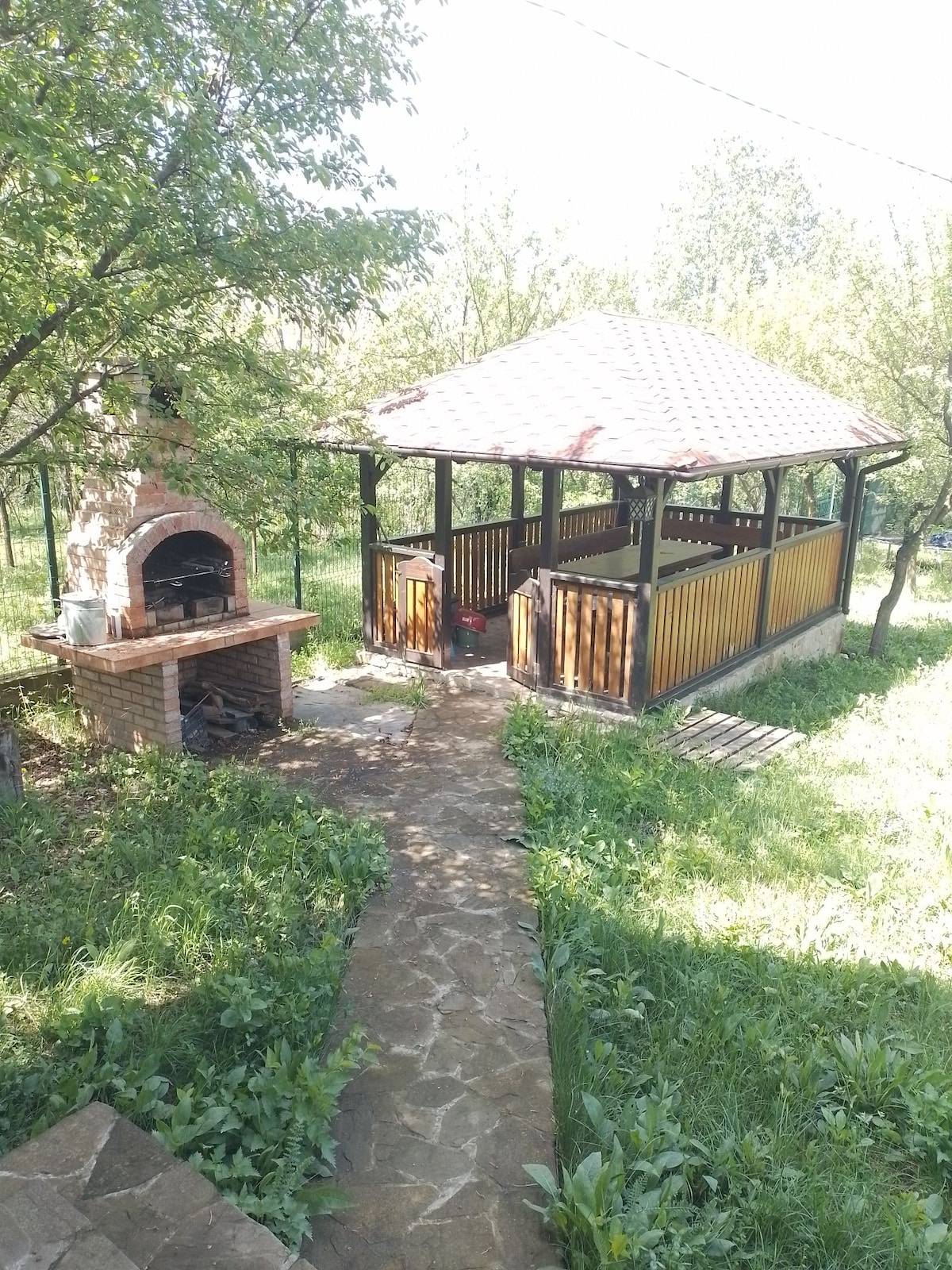 A charming outdoor gazebo is set within a lush garden, featuring wooden railings and a tiled roof. Adjacent to the gazebo, a stone barbecue grill offers a convenient cooking area. The pathway leading up is surrounded by green grass and trees, creating a serene outdoor space.