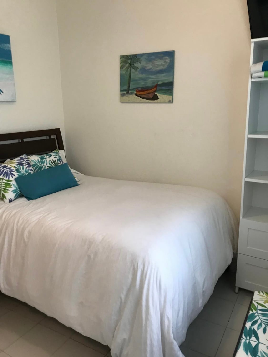 A bright sleeping area features a spacious bed adorned with white linens and a variety of colorful decorative pillows. A tropical-themed wall art piece hangs above, complementing the serene atmosphere. A tall shelving unit stands nearby, providing additional storage space.