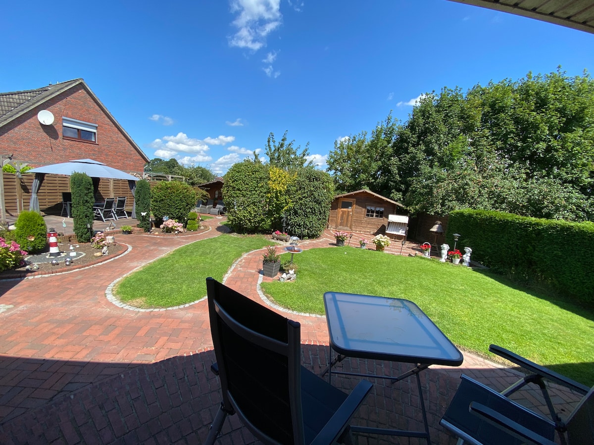 A well-maintained garden is shown, featuring a neatly paved path surrounded by green grass and various shrubs. Several garden structures, including a shed, are visible in the background, along with ornamental decorations throughout the space, creating an inviting outdoor area.