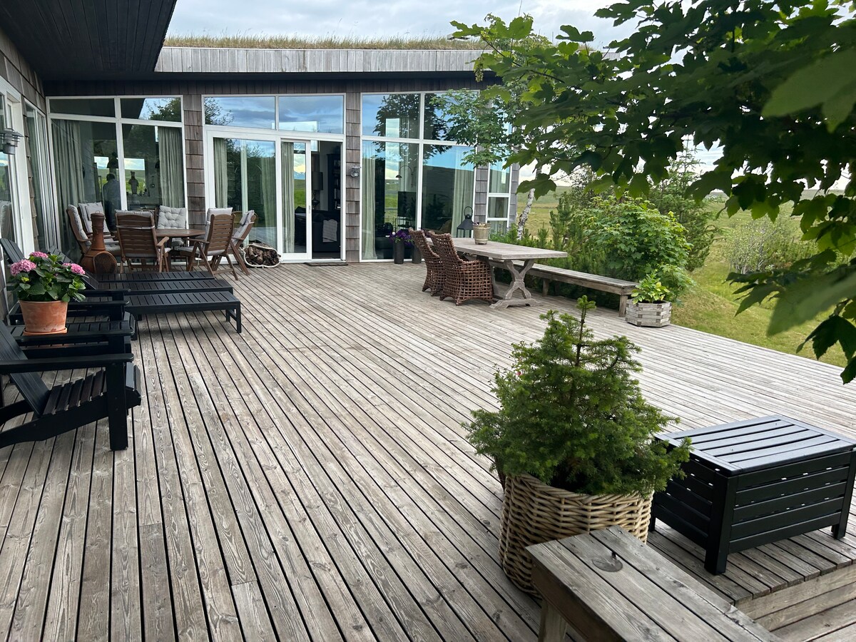 An outdoor patio features wooden decking, furnished with two lounge chairs and a dining table surrounded by various plants. Large glass doors provide access to the interior, while greenery and trees frame the outdoor space.