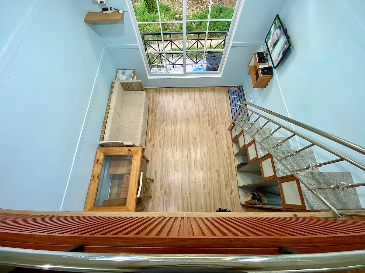 The living area is observed from above, featuring light blue walls and wooden flooring. A beige sofa and a glass coffee table are positioned on the right. A television is mounted on the wall opposite a large window, allowing natural light to fill the space.