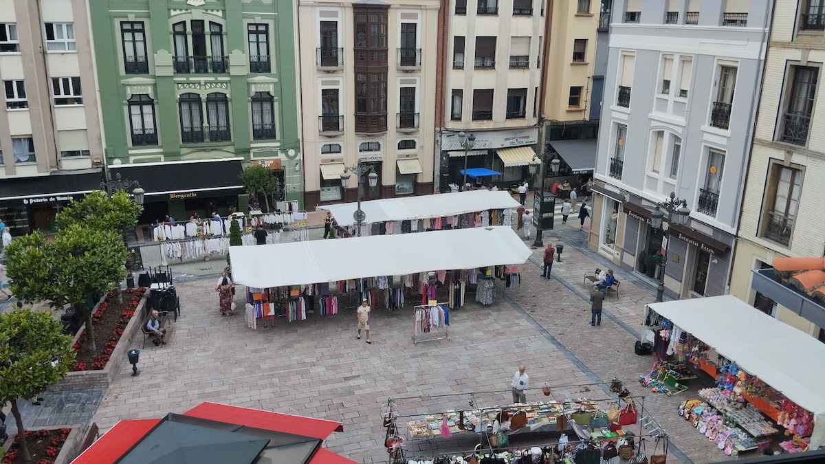 A lively market scene is captured from above, showcasing a spacious plaza filled with vendor stalls covered by white canopies. Colorful clothing and various goods are displayed along the cobblestone surface. Surrounding buildings feature a mix of architectural styles, contributing to the historic charm of the area.