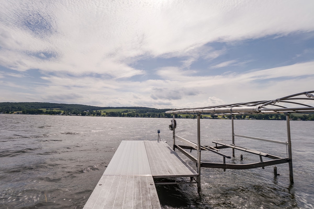 A spacious dock extends into the shimmering waters of Waneta Lake, framed by rolling green hills in the background. The sky features a blend of soft clouds and ample sunlight, reflecting upon the lake's surface, creating a serene waterfront setting.