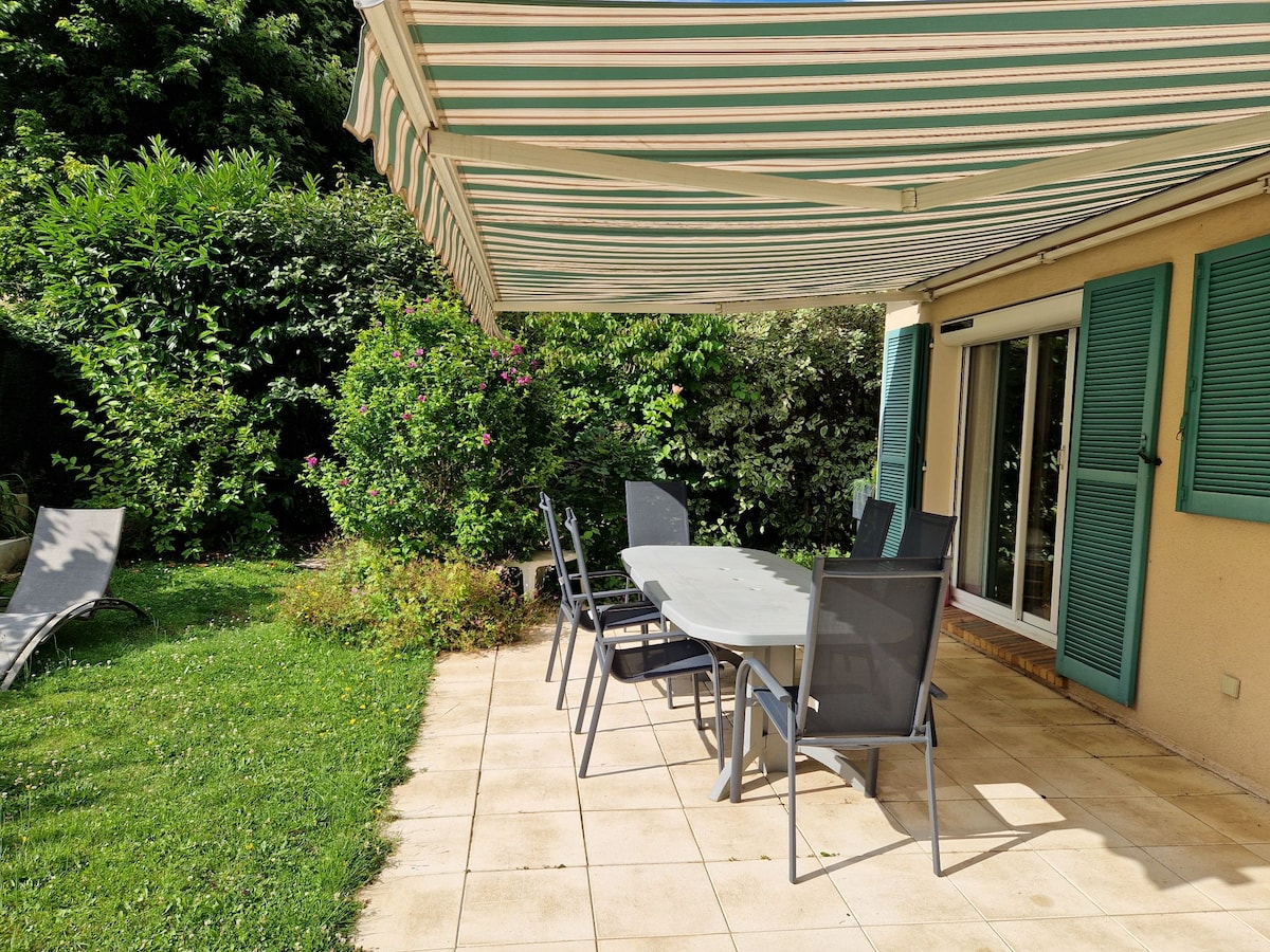 A terrace area is highlighted by a large table surrounded by six chairs, set against a backdrop of lush greenery. An awning provides shade, enhancing the space for outdoor dining or relaxation. Additional lounge chairs are positioned nearby on a tiled surface.
