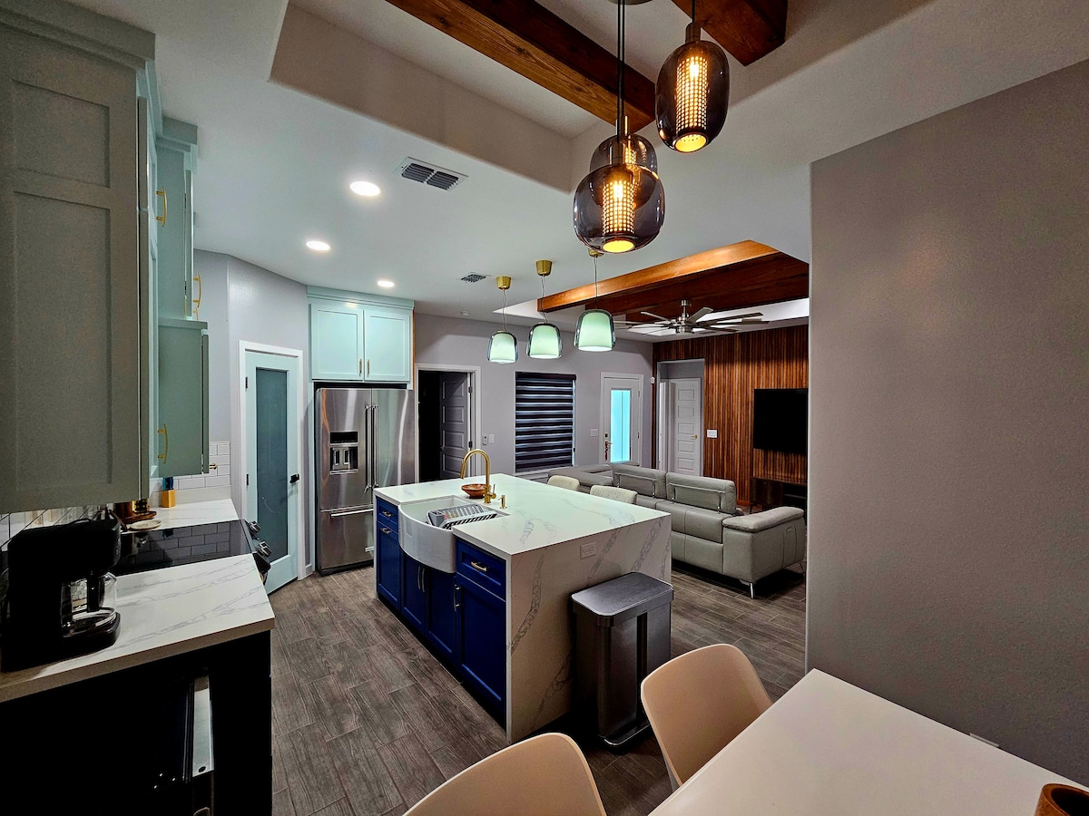 An open kitchen area features modern cabinetry and a large island with a sink. Overhead pendant lights provide ambient lighting. Comfortable seating is arranged in the adjoining living area, with a large sectional sofa facing a wall-mounted TV. Natural light enters through multiple windows.