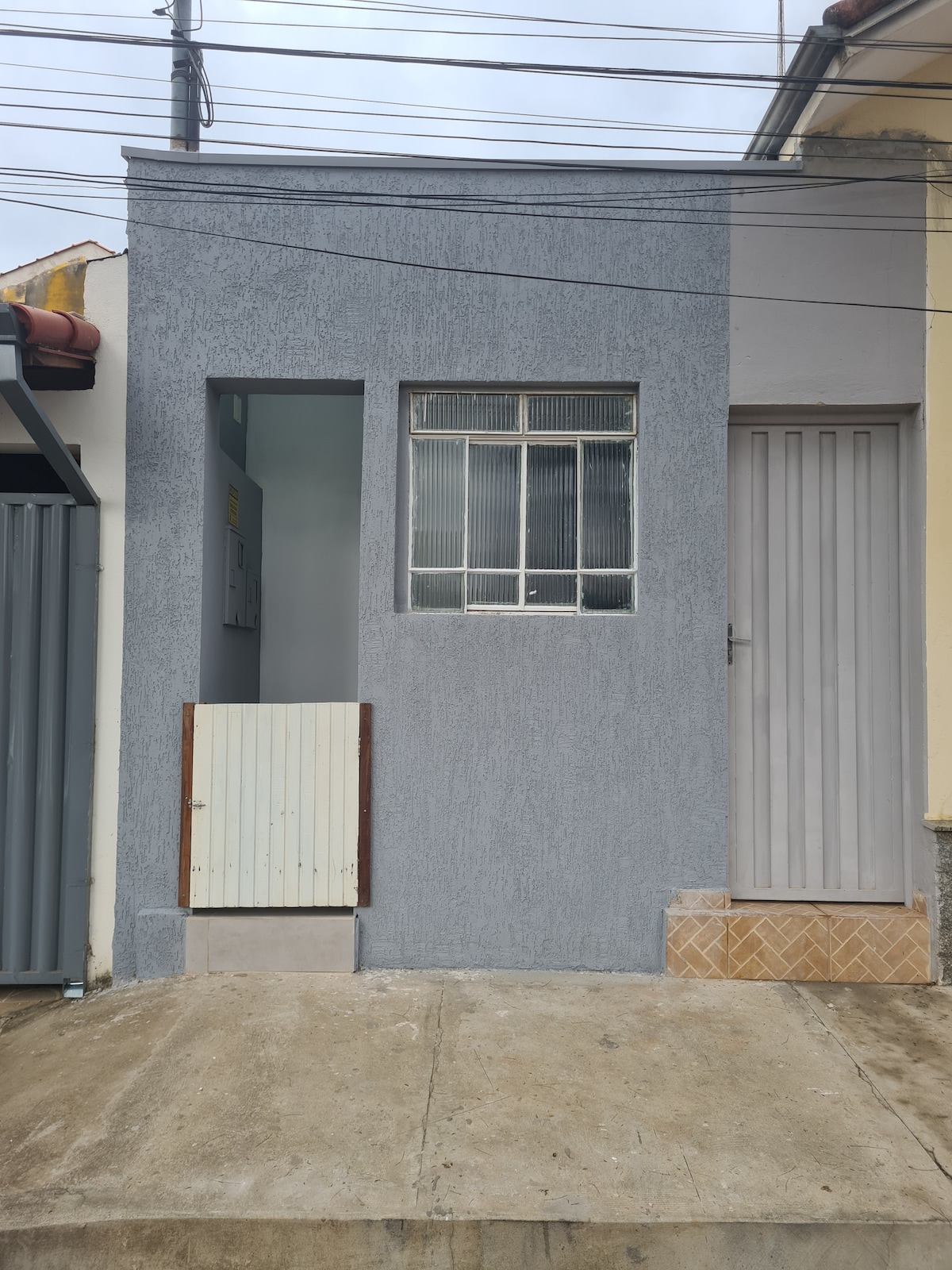 The exterior of a compact building presents a gray facade, featuring a large window with grid design. A small white door is positioned to the left, and a light-colored door is seen to the right. Electrical wires are visible overhead against the cloudy sky.