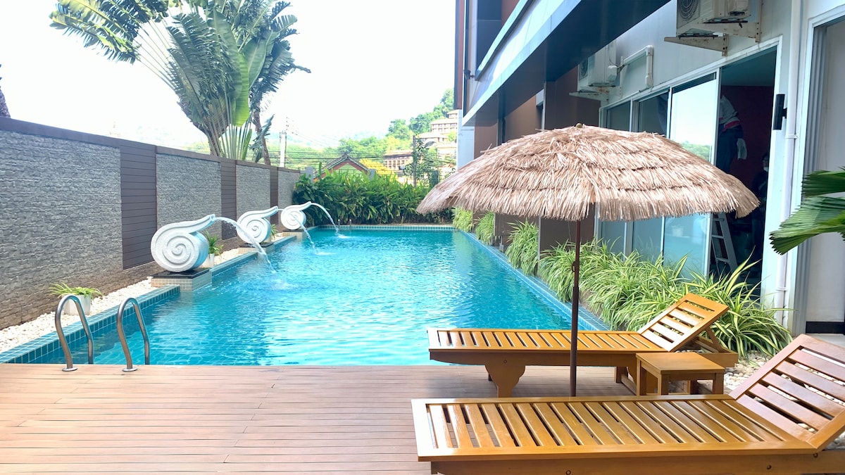 A serene pool area is captured, featuring a clear blue pool surrounded by lush greenery. Two wooden lounge chairs are positioned on a wooden deck, accompanied by a thatched umbrella for shade. The soothing sounds of water flow enhance the tranquil setting.