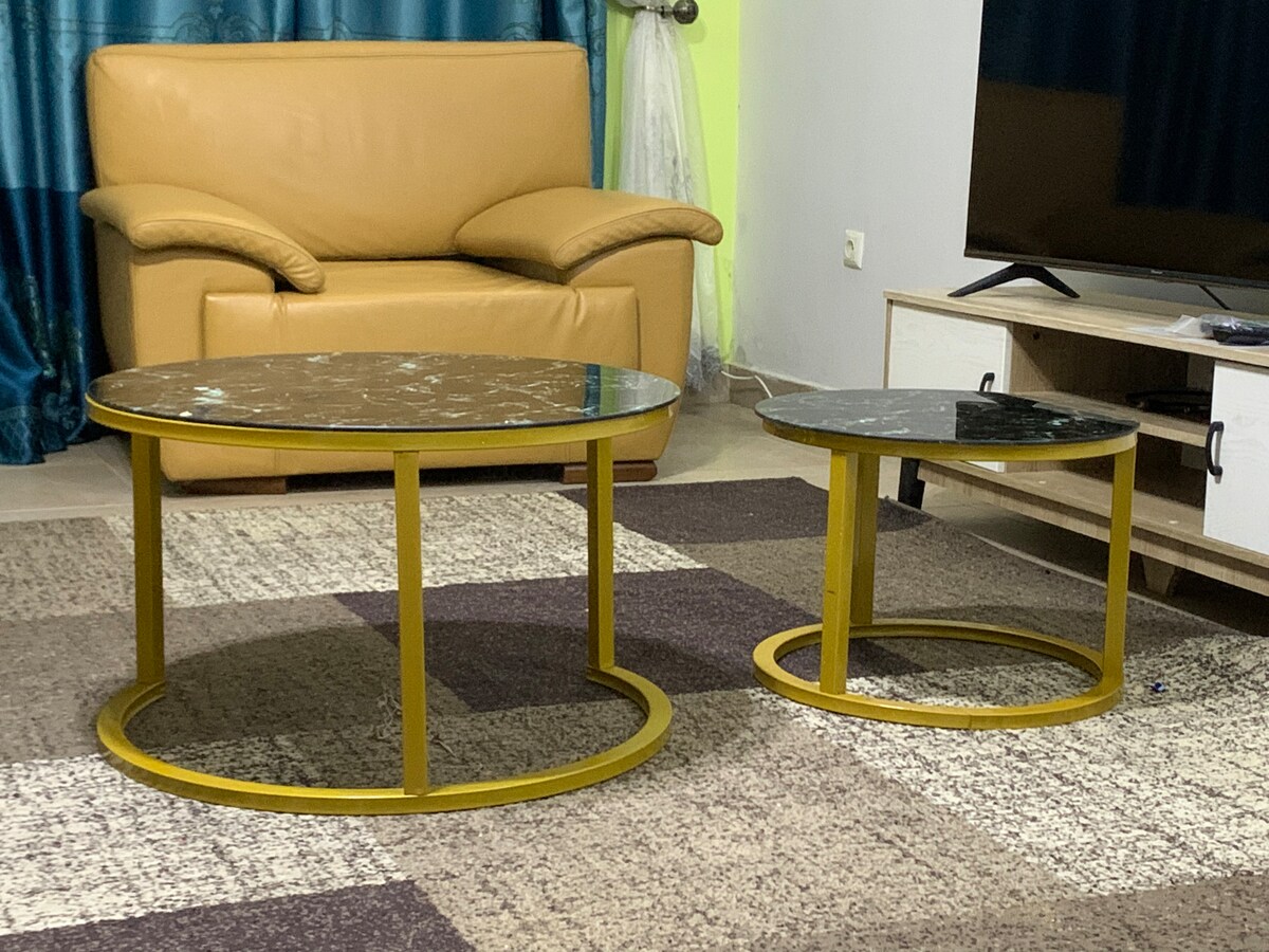 A living area is highlighted by two circular coffee tables, one large and one small, featuring marble-patterned tops and sleek yellow metal bases. A beige leather sofa provides comfortable seating in the background, complemented by a textured area rug in various neutral tones.