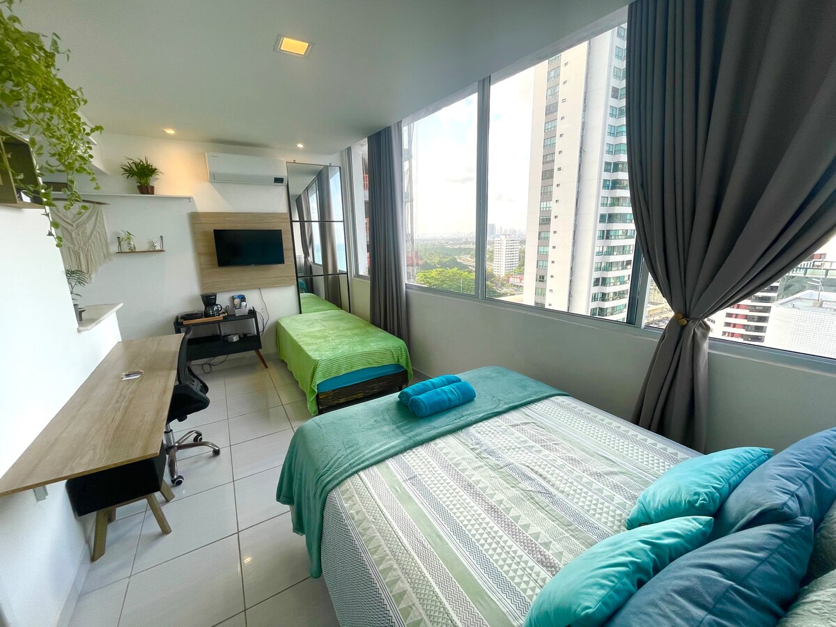 A bright and airy sleeping area features large windows providing abundant natural light and views of the cityscape. Two beds are presented with neatly arranged linens, and a workspace is equipped with a desk and a chair. Elegant curtains frame the windows, adding a touch of comfort.