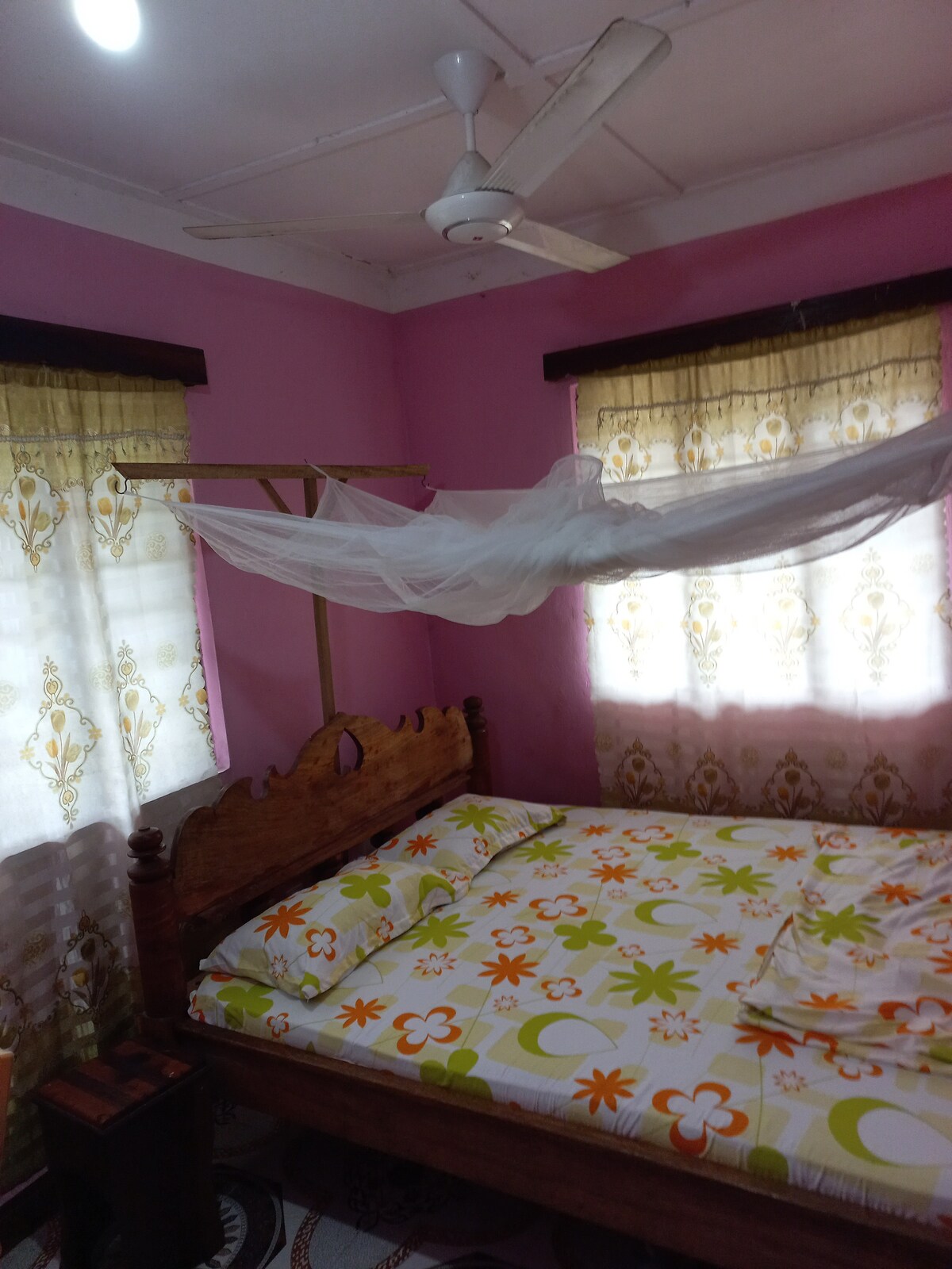 A cozy bedroom is presented, featuring a wooden bed adorned with a vibrant floral-patterned sheet. Soft, natural light filters through two window coverings, while a mosquito net hangs over the bed, enhancing comfort and tranquility in the space.