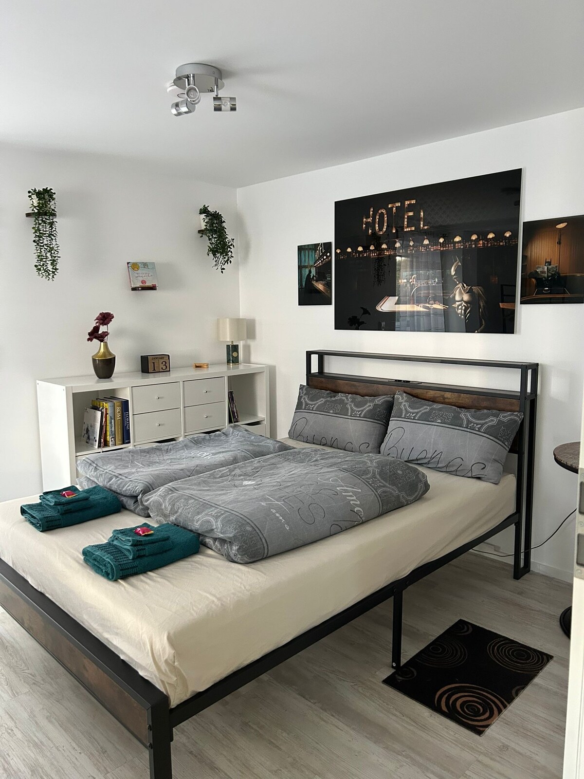 A cozy bedroom features a spacious bed with patterned bedding and two sets of neatly arranged towels at the foot. A wall-mounted photo reflects a hotel ambiance, while shelves and a dresser provide storage space. Indoor plants add a touch of green to the decor.