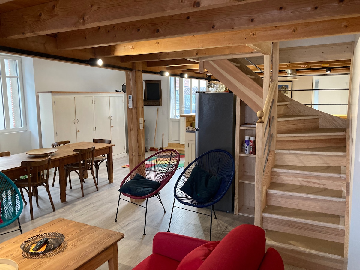 An open living area features wooden beams and a staircase leading to the mezzanine. A dining table is visible alongside chairs, and two colorful lounge chairs are arranged nearby. A television is mounted on the wall, complementing the cozy ambiance.