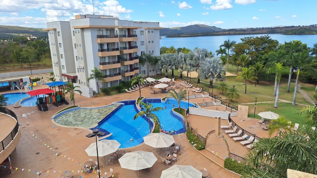 A vast pool area is framed by palm trees and lounge chairs, offering relaxation by the water. The hotel building stands nearby, featuring multiple floors and large windows. The serene background shows the expansive Lake Corumbá, reflecting the sky and surrounded by lush greenery.
