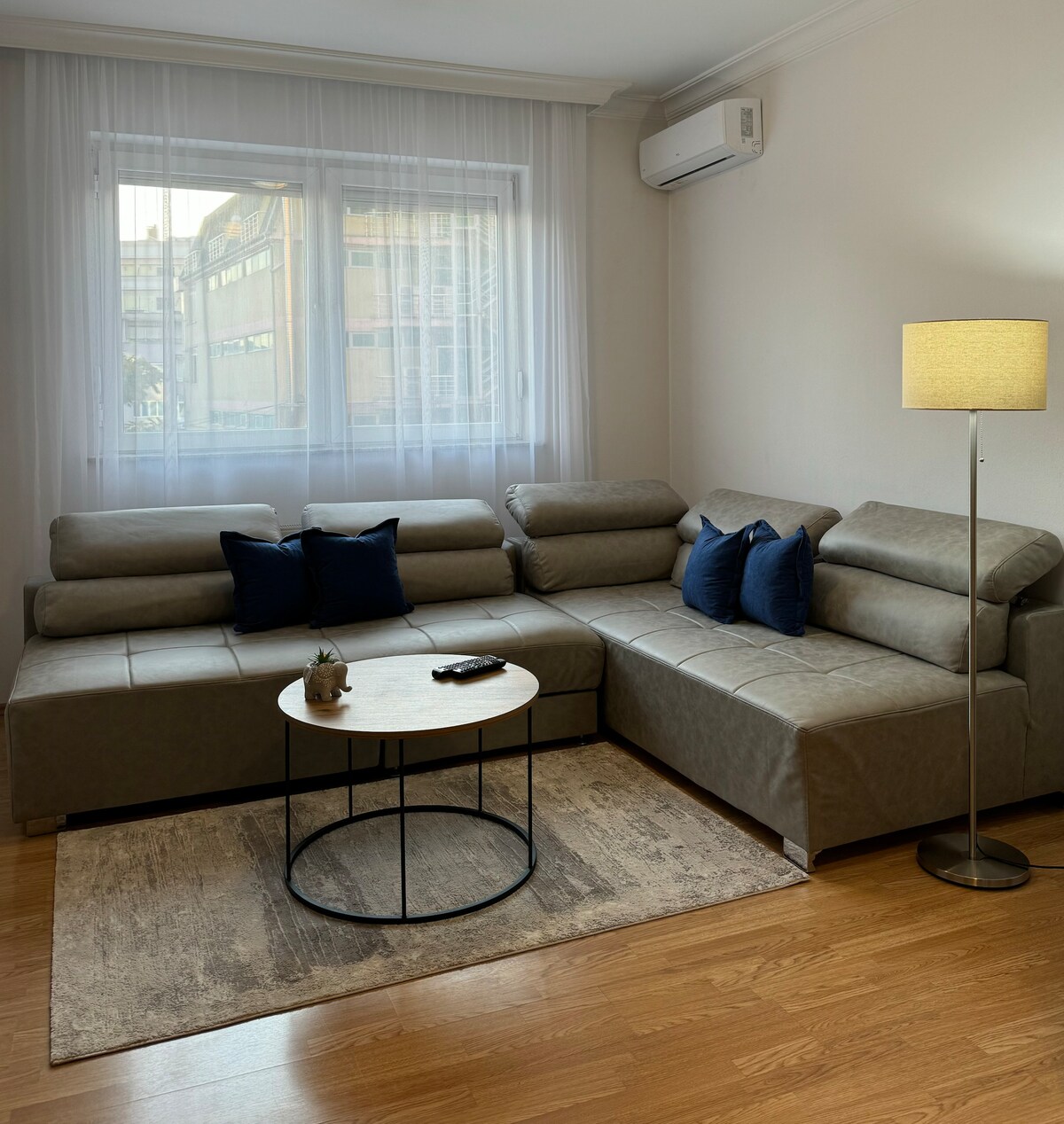A comfortable living area features a sectional sofa with multiple cushions, complemented by a round coffee table positioned on a soft area rug. Natural light enters through a large window adorned with sheer curtains, and a standing lamp provides additional lighting.
