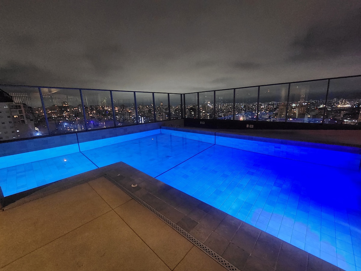 A rooftop pool with a luminous blue hue is positioned against a backdrop of city lights. The pool area features a surrounding glass railing, providing an unobstructed view of the skyline. The deck is constructed with smooth tiles, contributing to a modern atmosphere.