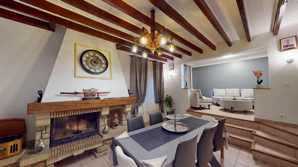 A spacious living area features a rustic fireplace with decorative items above. A large dining table with gray chairs is positioned in front of a large window. An inviting sofa area is elevated, with soft lighting and a wall adorned with subtle decor, creating a cozy ambiance.