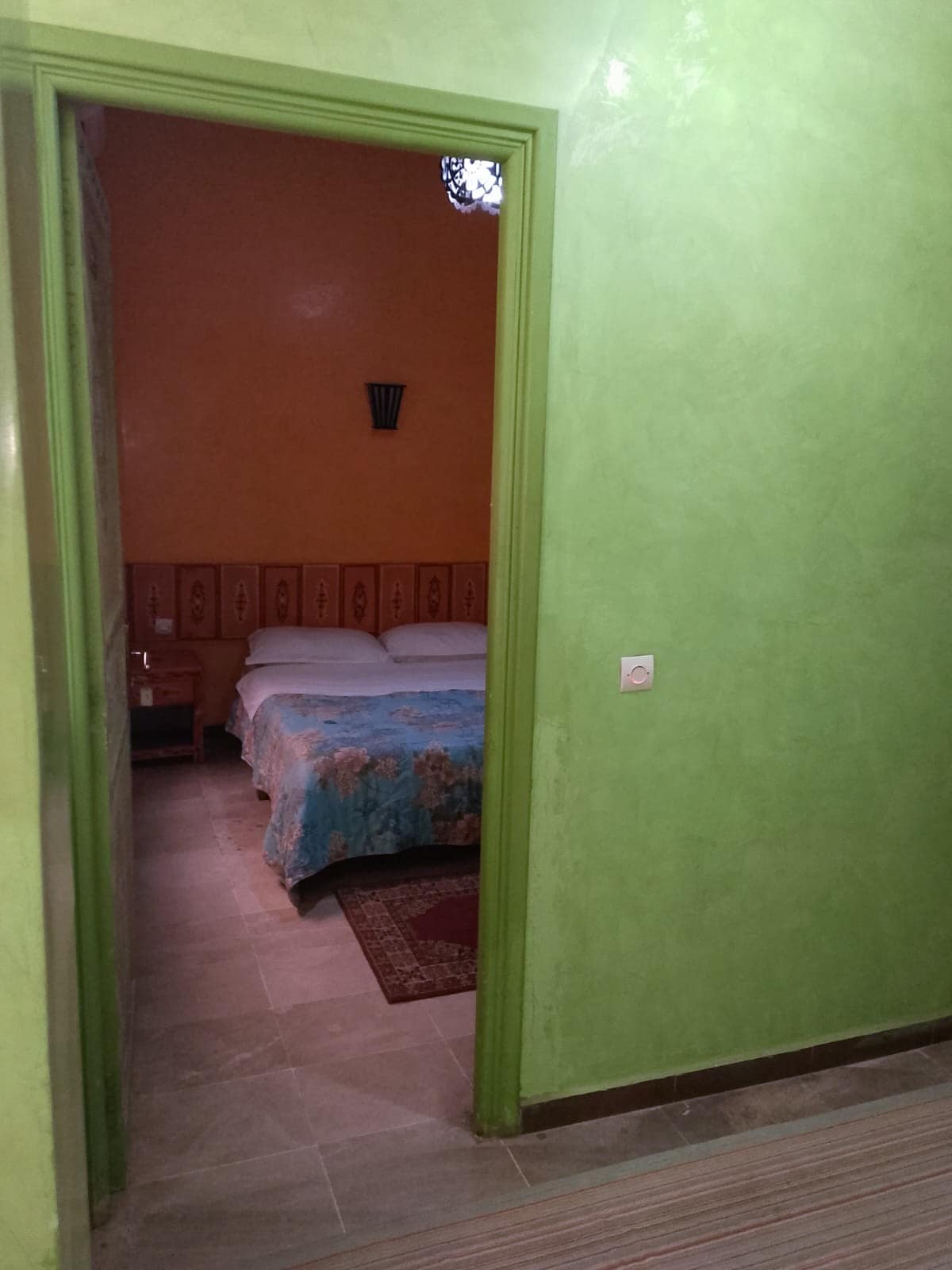 A vibrant entrance leads into a room featuring a spacious bed with a blue coverlet. The walls are painted in warm orange and green tones, complemented by decorative wooden paneling. A rug adds warmth to the tiled floor, while a lantern provides soft lighting.