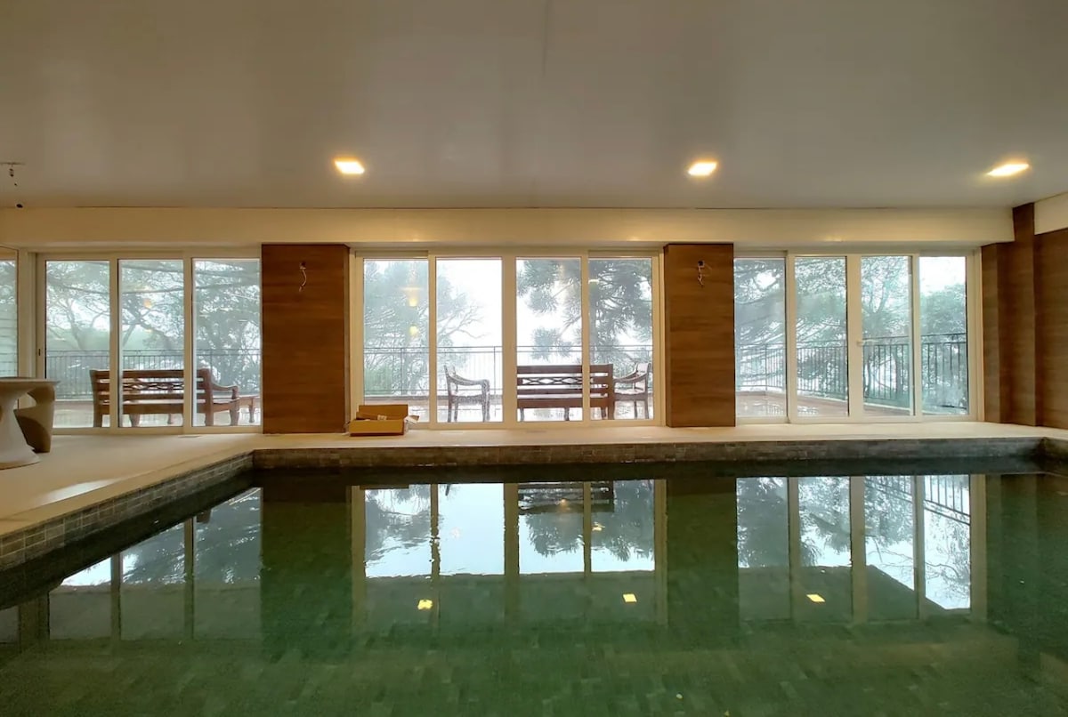 An indoor pool area is highlighted by large floor-to-ceiling windows, allowing soft natural light to fill the space. Comfortable seating is positioned nearby, overlooking the serene exterior landscape, creating a calm and relaxing environment.