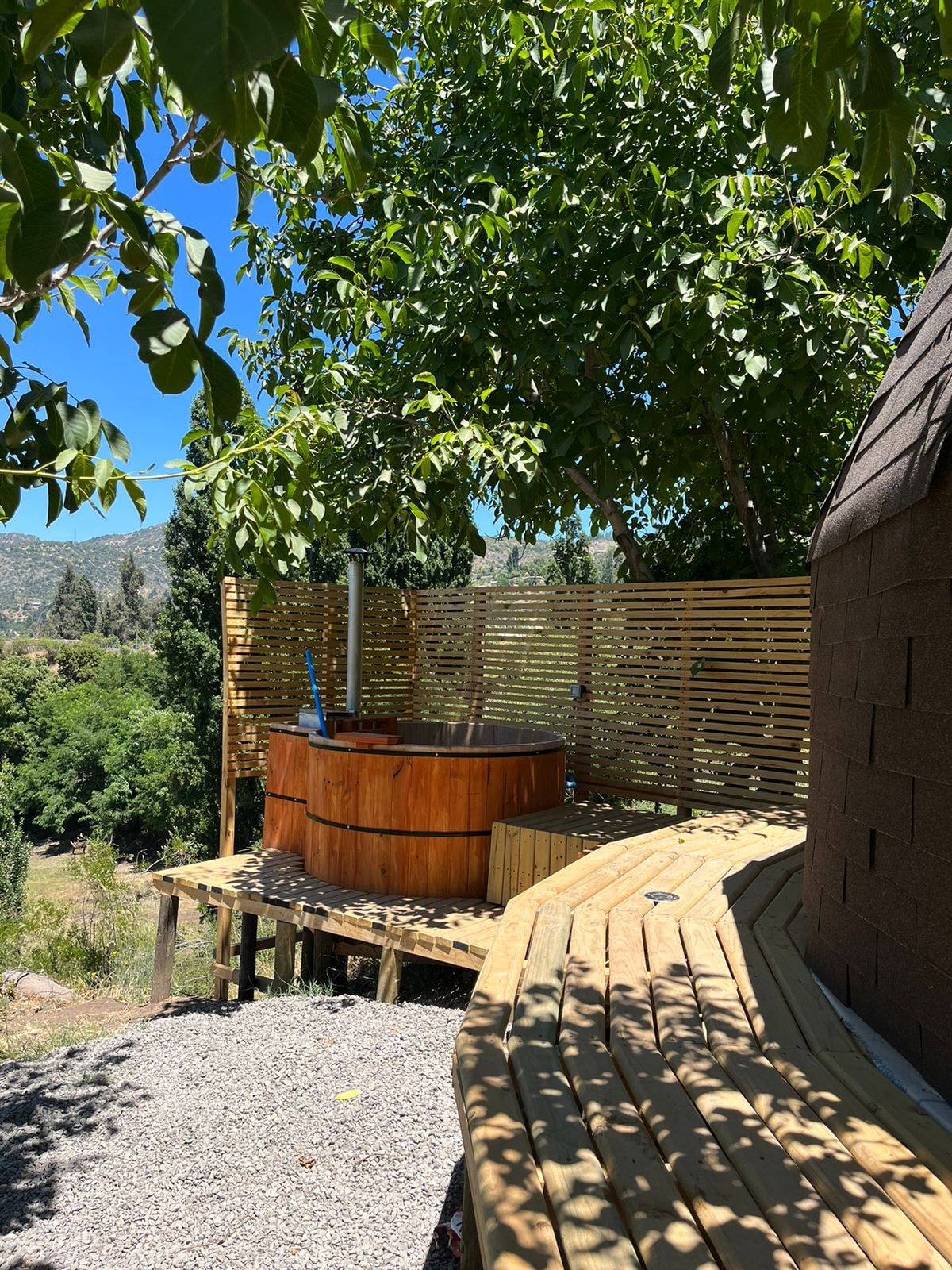 A wooden hot tub is set on a raised deck surrounded by greenery, offering a relaxed outdoor atmosphere. A light-toned wooden walkway curves gently around the area, enhancing the connection with nature. The scene is framed by leafy trees, providing shade and a sense of privacy.