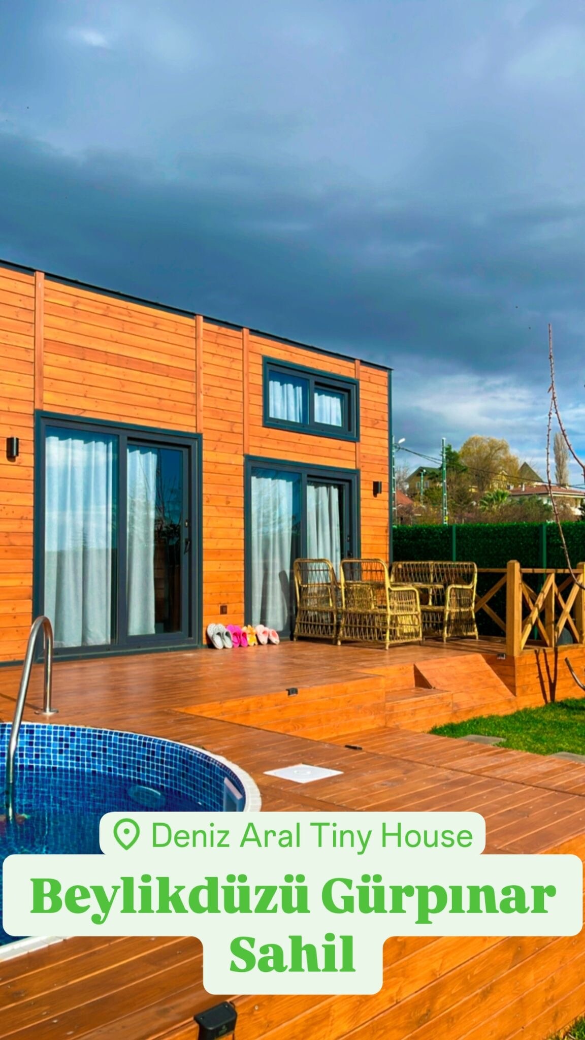 A wooden exterior of a tiny house is shown, featuring large windows and a cozy seating area on the deck. A private hot tub is positioned nearby, surrounded by greenery. The sky is overcast, providing a serene backdrop to the modern design.